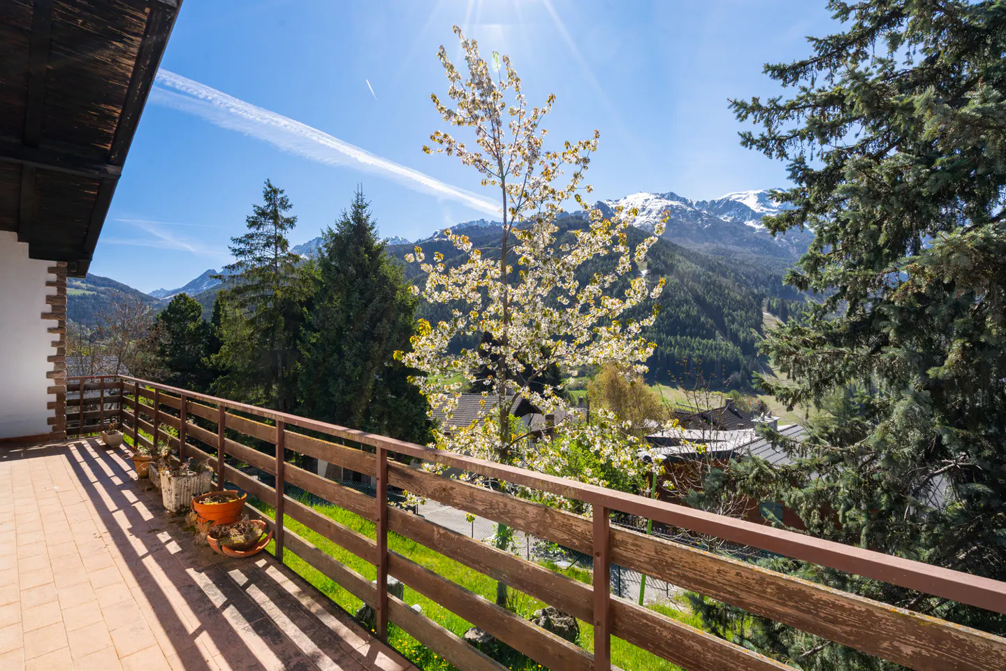 Balcony view with wooden railing, potted plants, and mountain backdrop under a sunny blue sky.