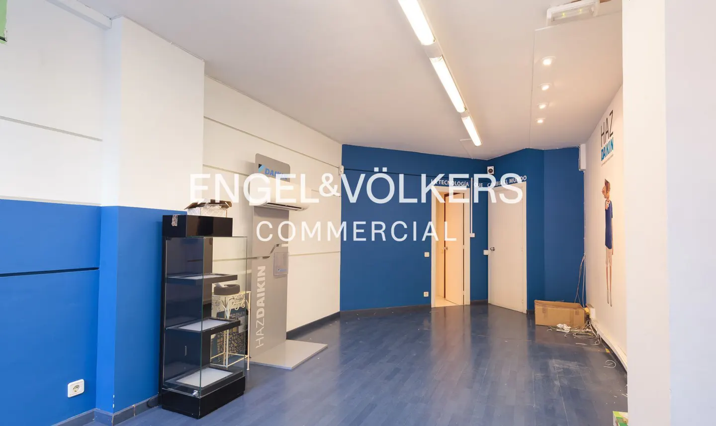 Empty commercial space with blue walls, a display case, and a door leading to another room. Fluorescent lights illuminate the space.