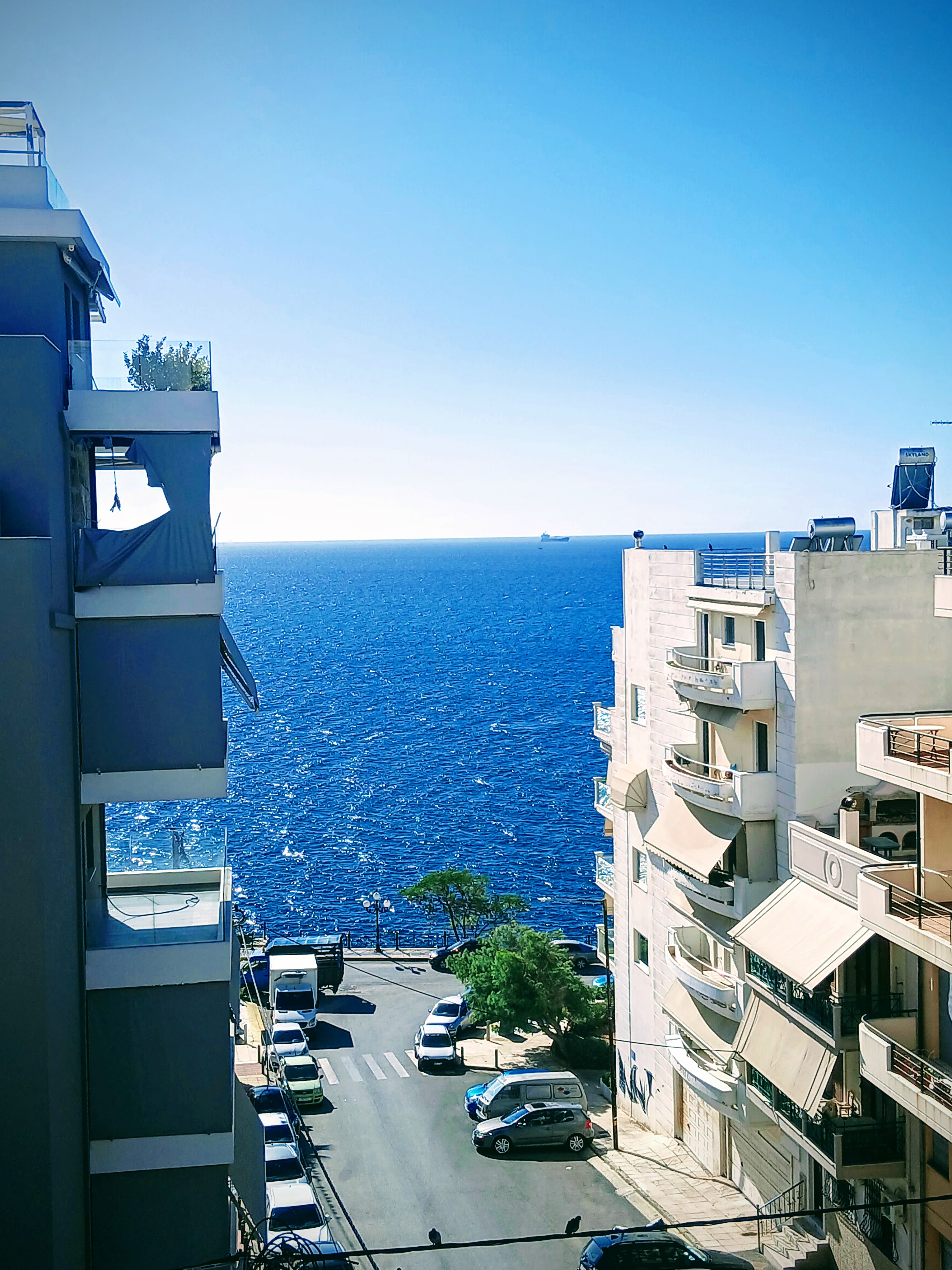 View of a street with parked cars between buildings, leading to a blue ocean under a clear sky.