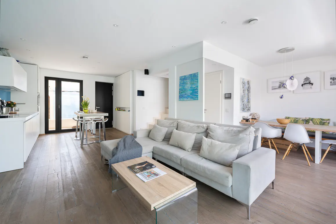 Bright, open-concept living space with a gray sofa, wood floors, and a dining area. Art adorns the white walls.