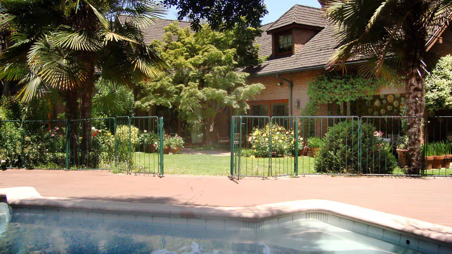 A backyard view of a brick house with a pool, green fence, trees, and flowers.