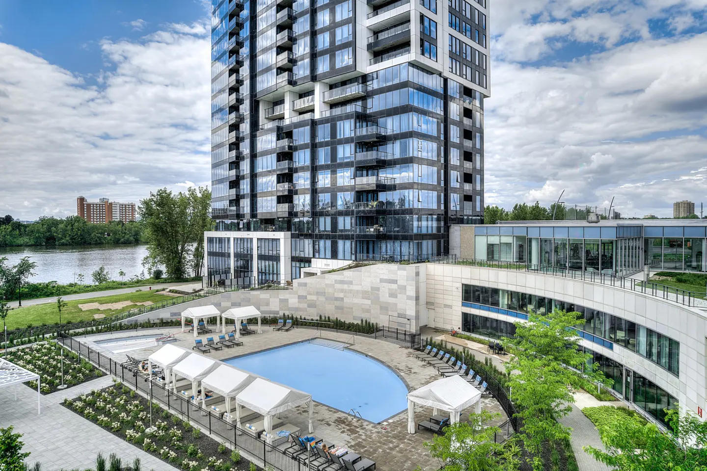 View of a modern high-rise condo with a pool, cabanas, and lounge chairs. A river and trees are in the background.
