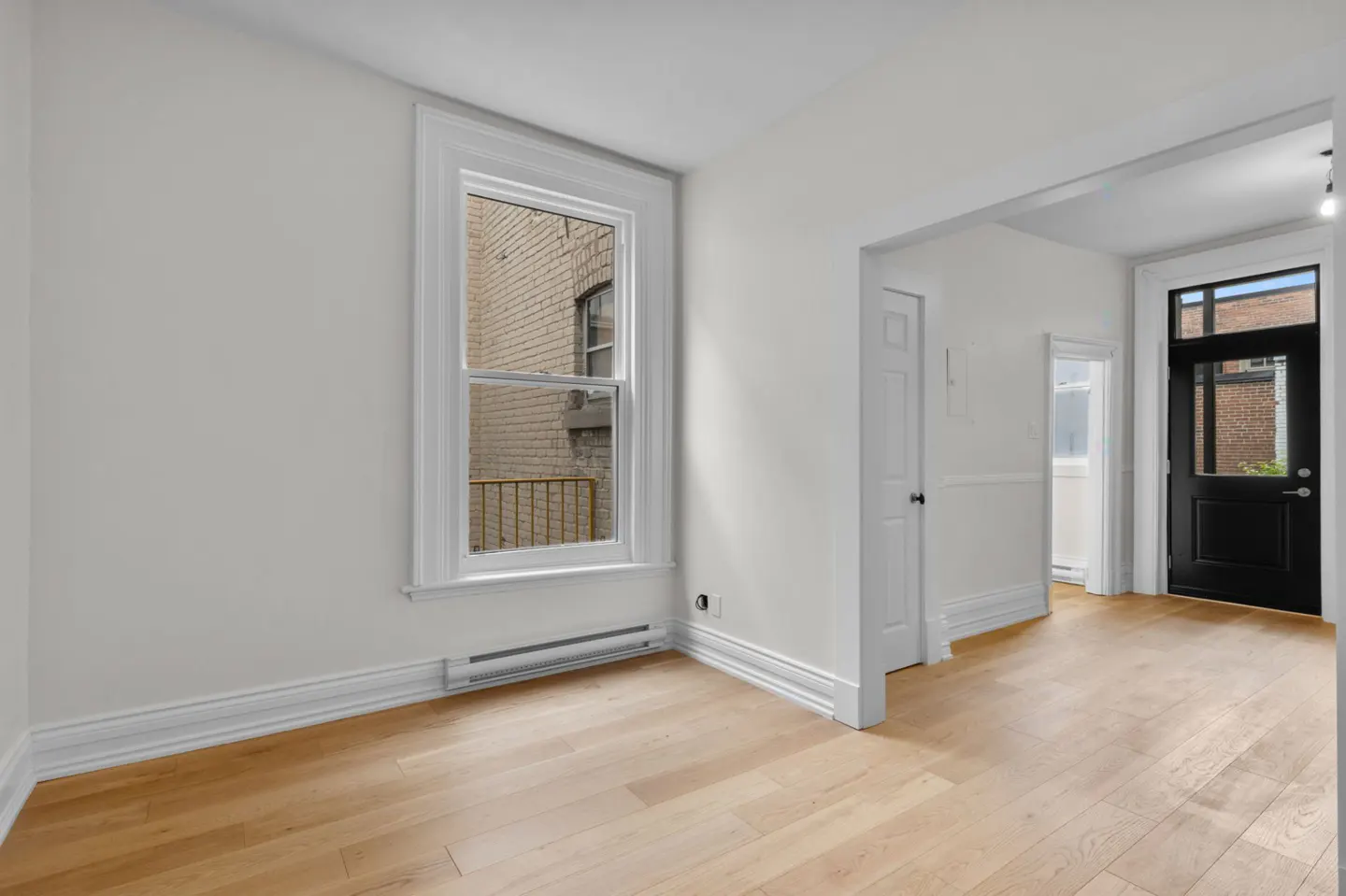 Bright, empty room with light wood floors, white walls, and trim. A window shows a brick building. A black door leads outside.