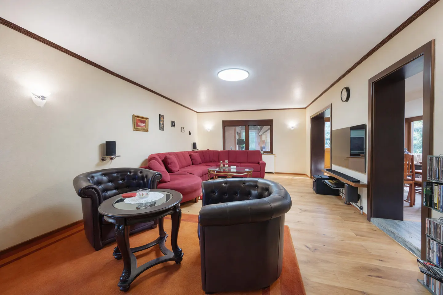 Living room with a red sectional sofa, brown leather chairs, and a dark wood table on an orange rug. A TV is mounted on the wall.