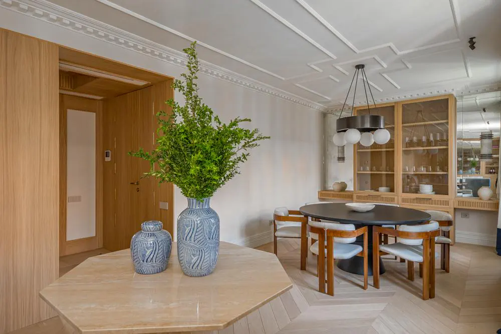 A dining room with light wood floors, a black oval table with chairs, and a blue vase with greenery on a marble table.