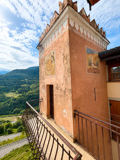 Exterior view of a peach-colored castle tower with a balcony overlooking a green valley and blue sky. The tower has a sundial and a painting on its walls.