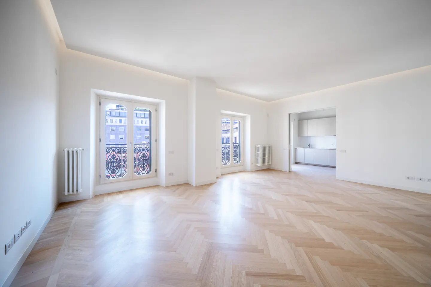 Bright, empty room with herringbone wood floors, white walls, and two windows with decorative ironwork. A kitchen is visible in the background.