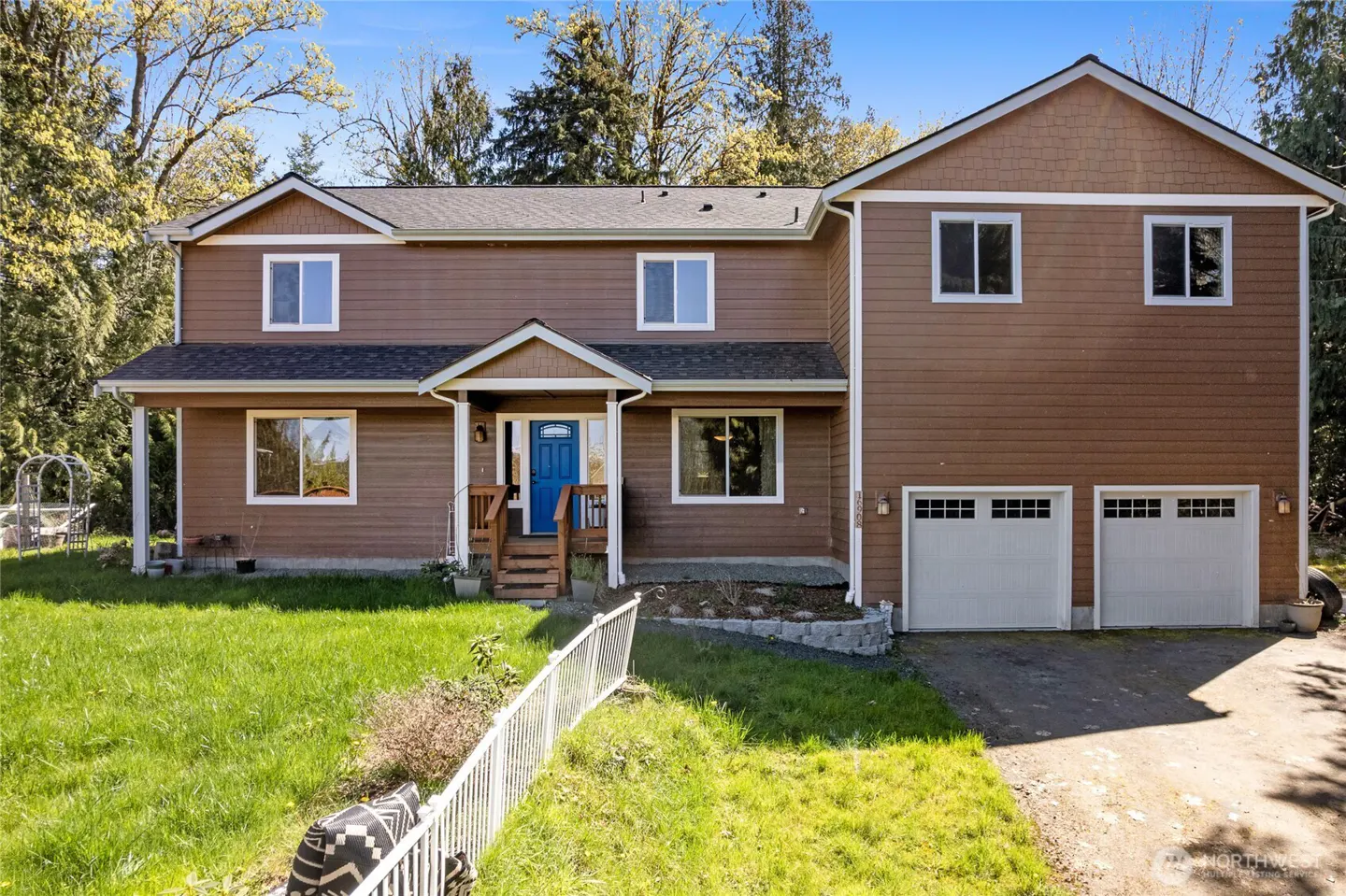 Two-story brown house with white trim, a blue front door, and a two-car garage. Green lawn and trees surround the house.