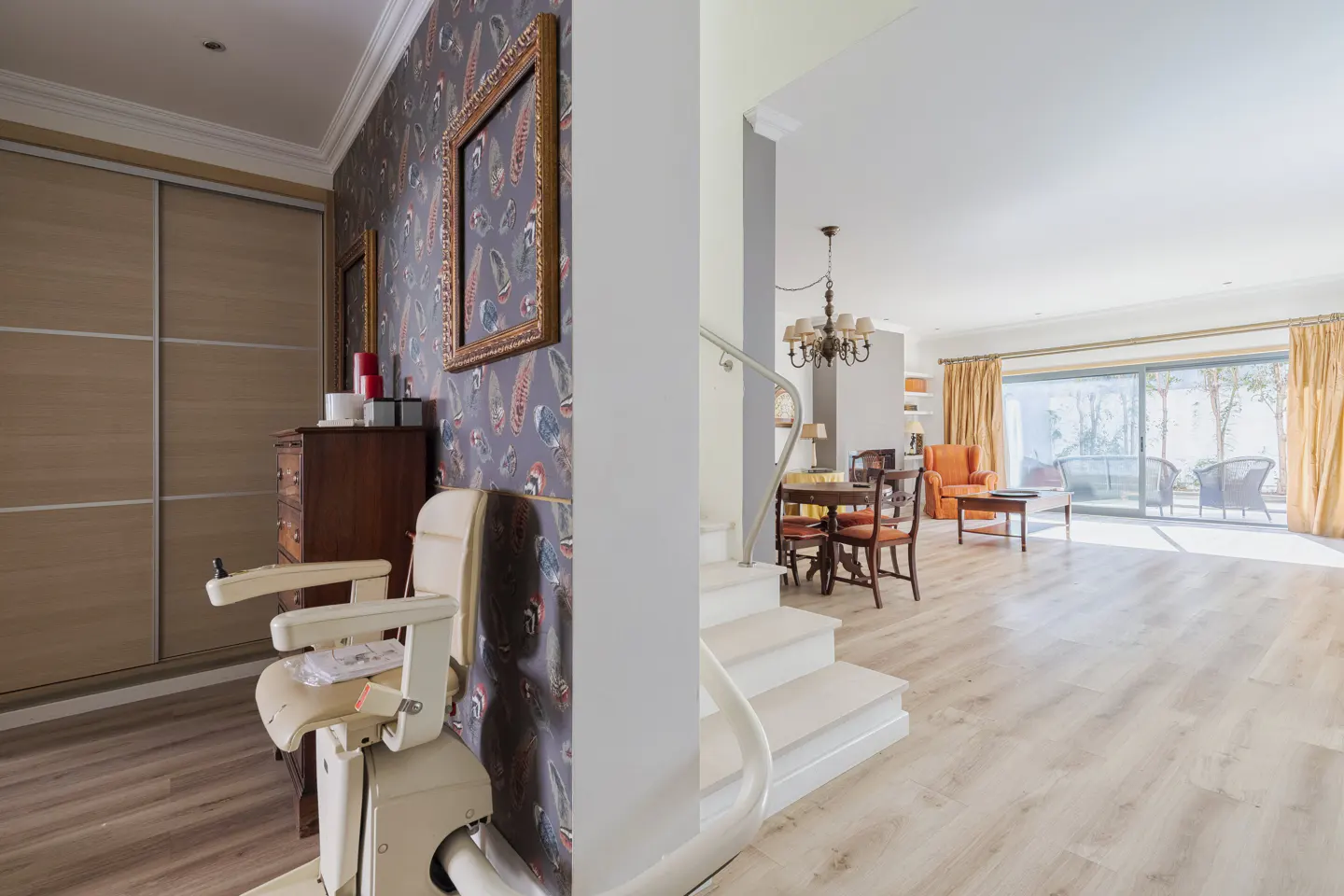 A stairlift sits in a hallway with feather-patterned wallpaper, leading to an open-plan living area with dining furniture.