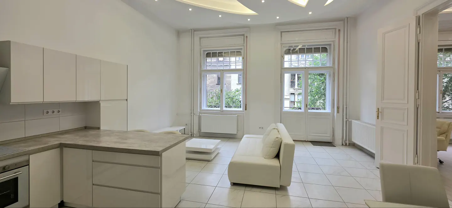 Modern white kitchen and living area with a white sofa, coffee table, and tall windows overlooking greenery.