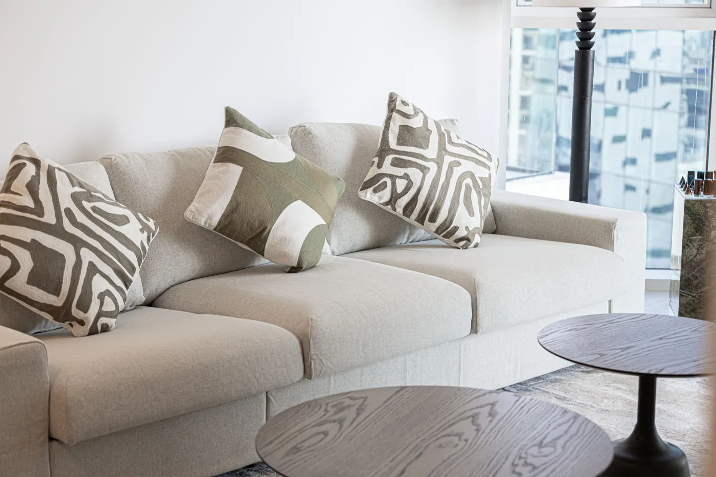 Living room with a light gray sofa, patterned pillows, and round wooden coffee tables. A window shows a city view.