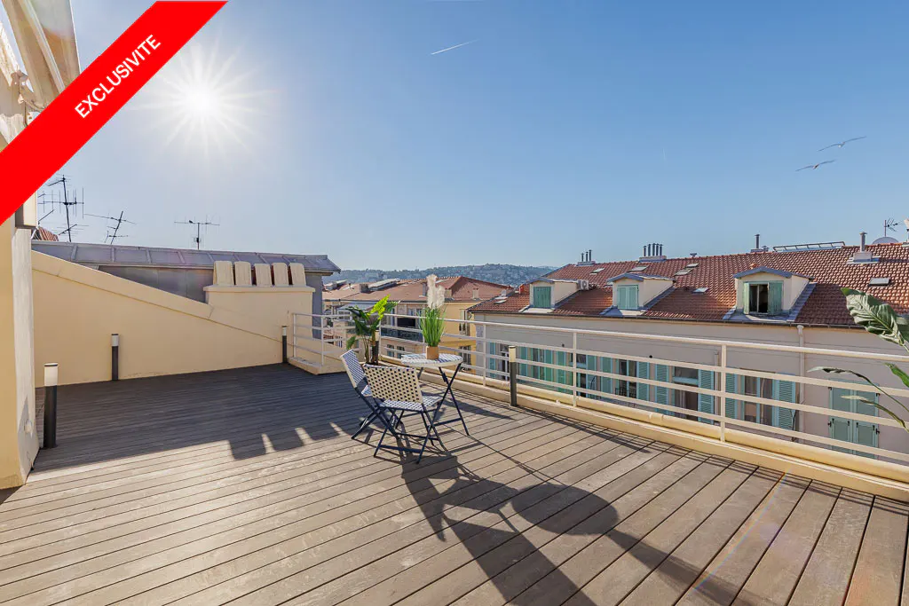 A rooftop patio with a table and chairs overlooks a city with red-tiled roofs under a clear blue sky. "Exclusive" banner in the corner.