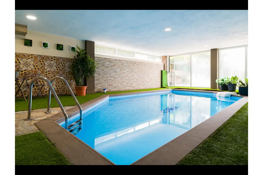 Indoor pool with blue water, surrounded by artificial grass and plants. Ladder for access. Tiled and brick walls.