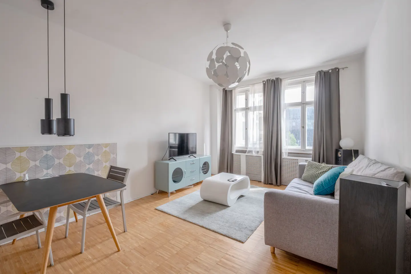 Bright living room with wood floors, a gray sofa with blue pillows, a blue cabinet with a TV, and a black table with chairs.