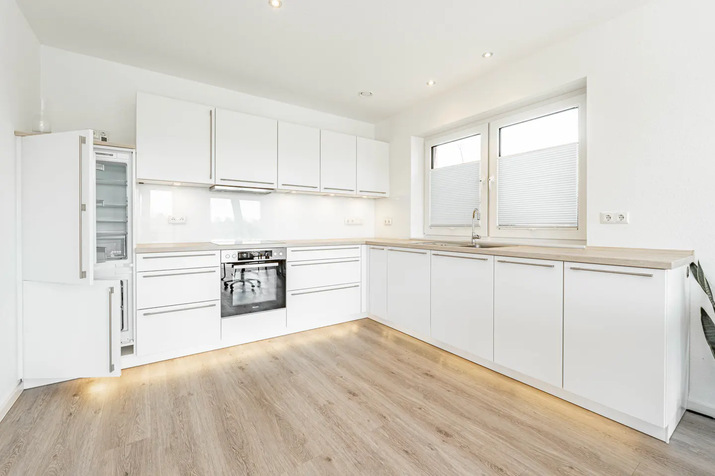 Bright, modern kitchen with white cabinets, light wood floors, and stainless steel appliances. A window with blinds lets in natural light.