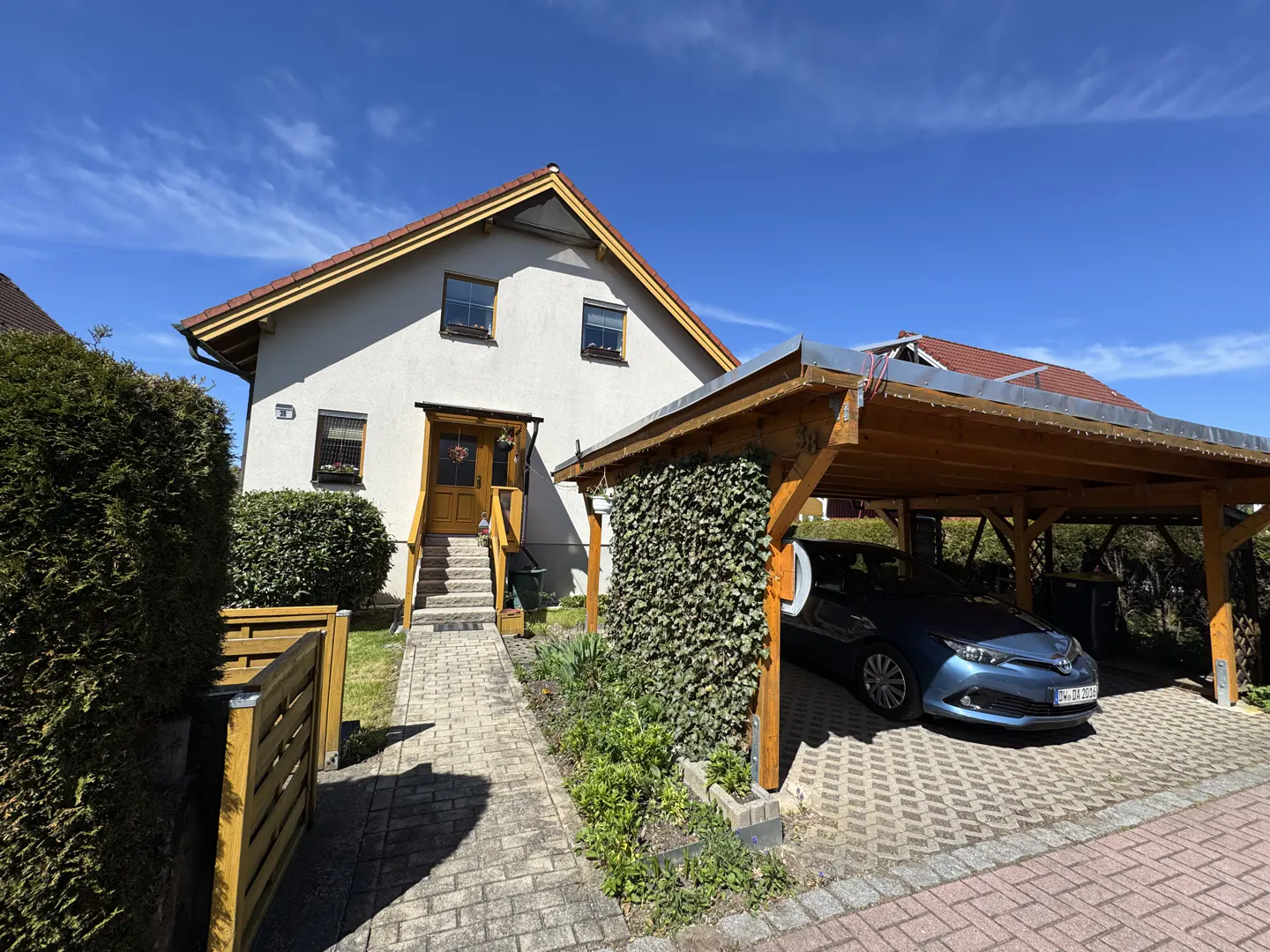 A two-story house with a brown roof and a blue car parked under a wooden carport.