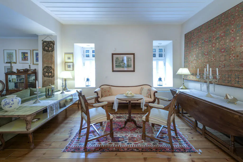 Living room with hardwood floors, white walls, and antique furniture. A patterned rug sits under a small table and chairs. Two windows flank a framed picture.