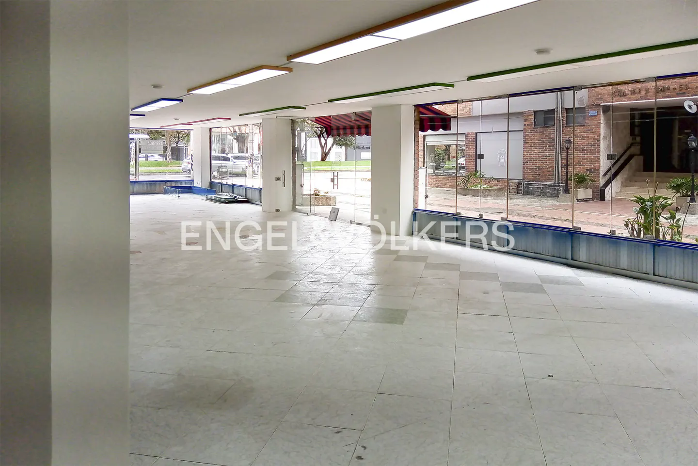Empty commercial space with white tile floor, large windows, and fluorescent lighting. Outside, a brick building and greenery are visible.