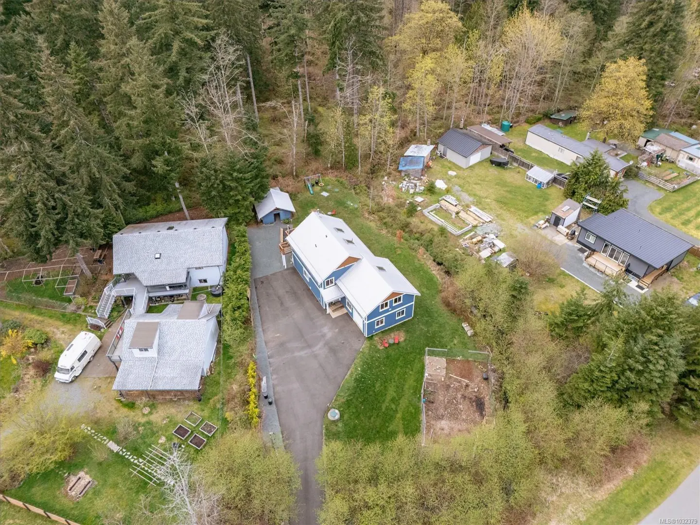 Aerial view of a blue house with a white roof, surrounded by trees and other houses in a rural setting.