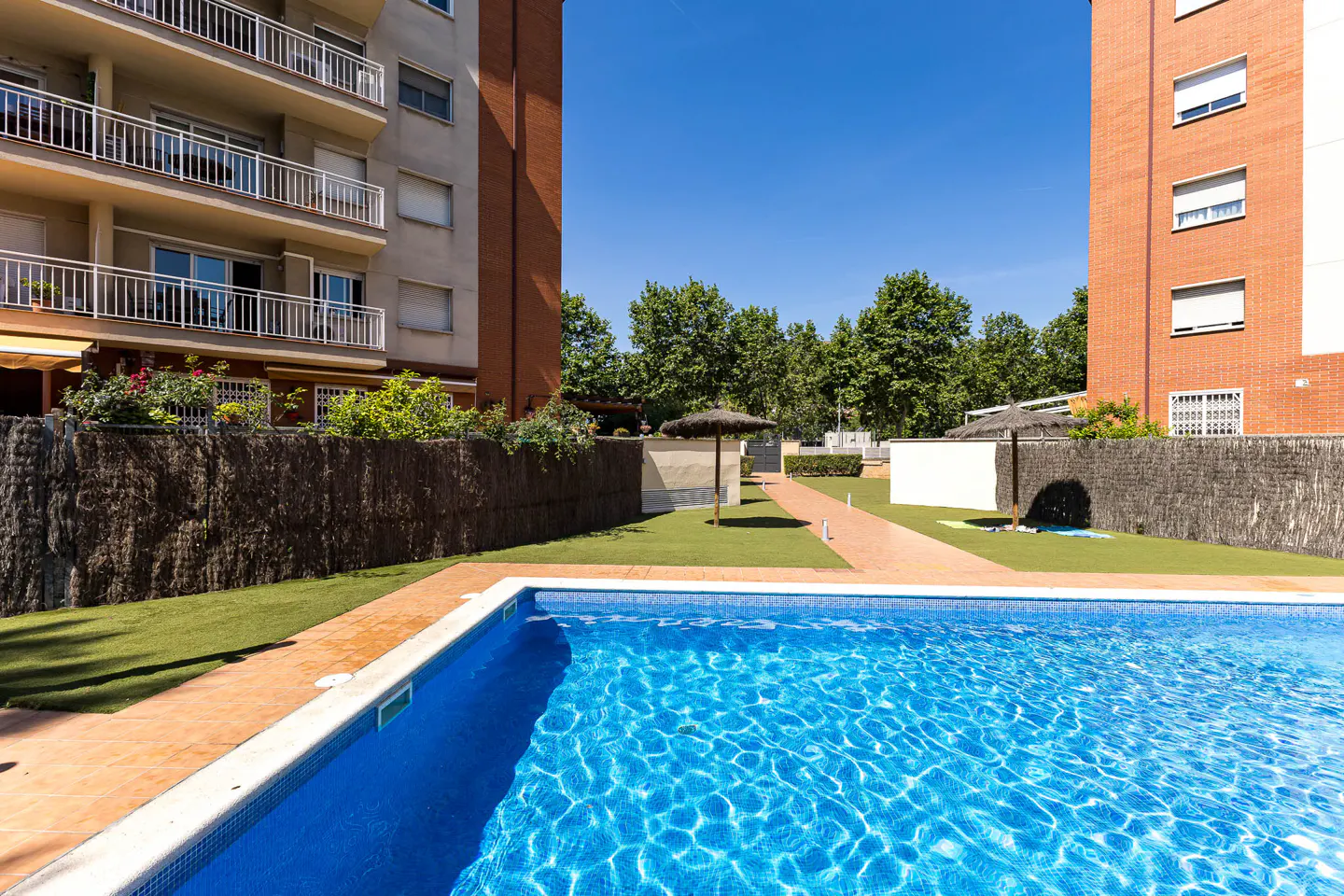 Outdoor pool with blue water, surrounded by grass and buildings under a clear blue sky.