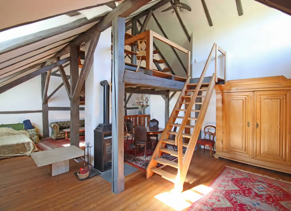 Attic living space with exposed beams, wood floors, and a loft area accessed by wooden stairs. A wood-burning stove and antique furniture are visible.