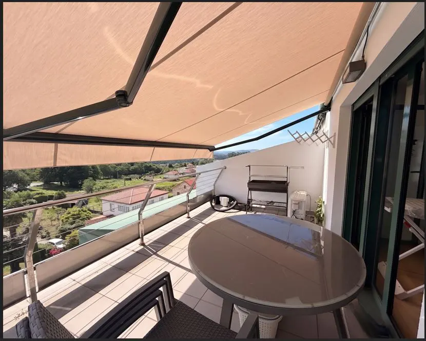 Balcony with a beige awning, round table, chairs, and a grill. A scenic view of houses and greenery is visible in the background.