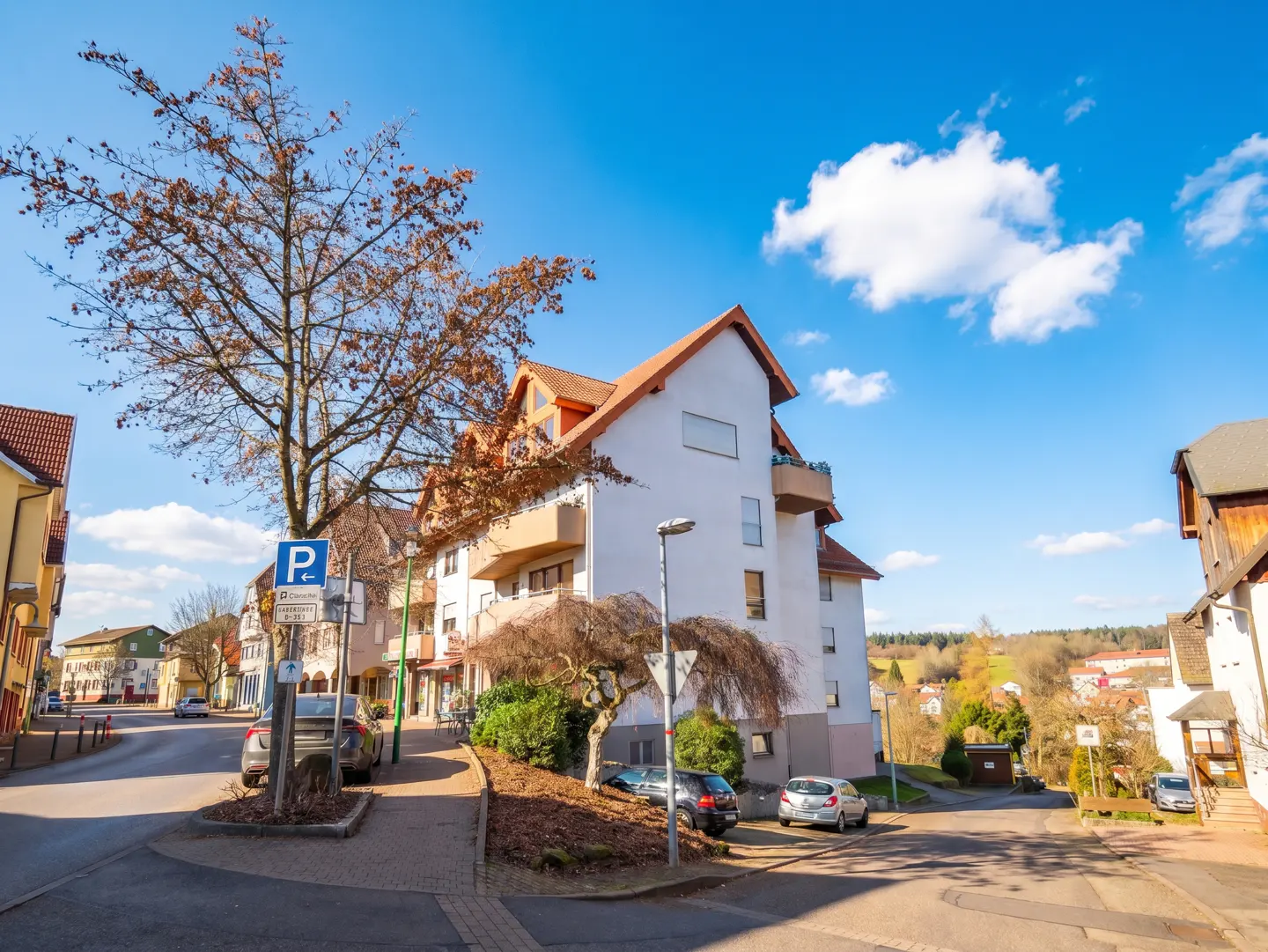 Street view of a white apartment building with a red tile roof on a sunny day with blue sky and white clouds.