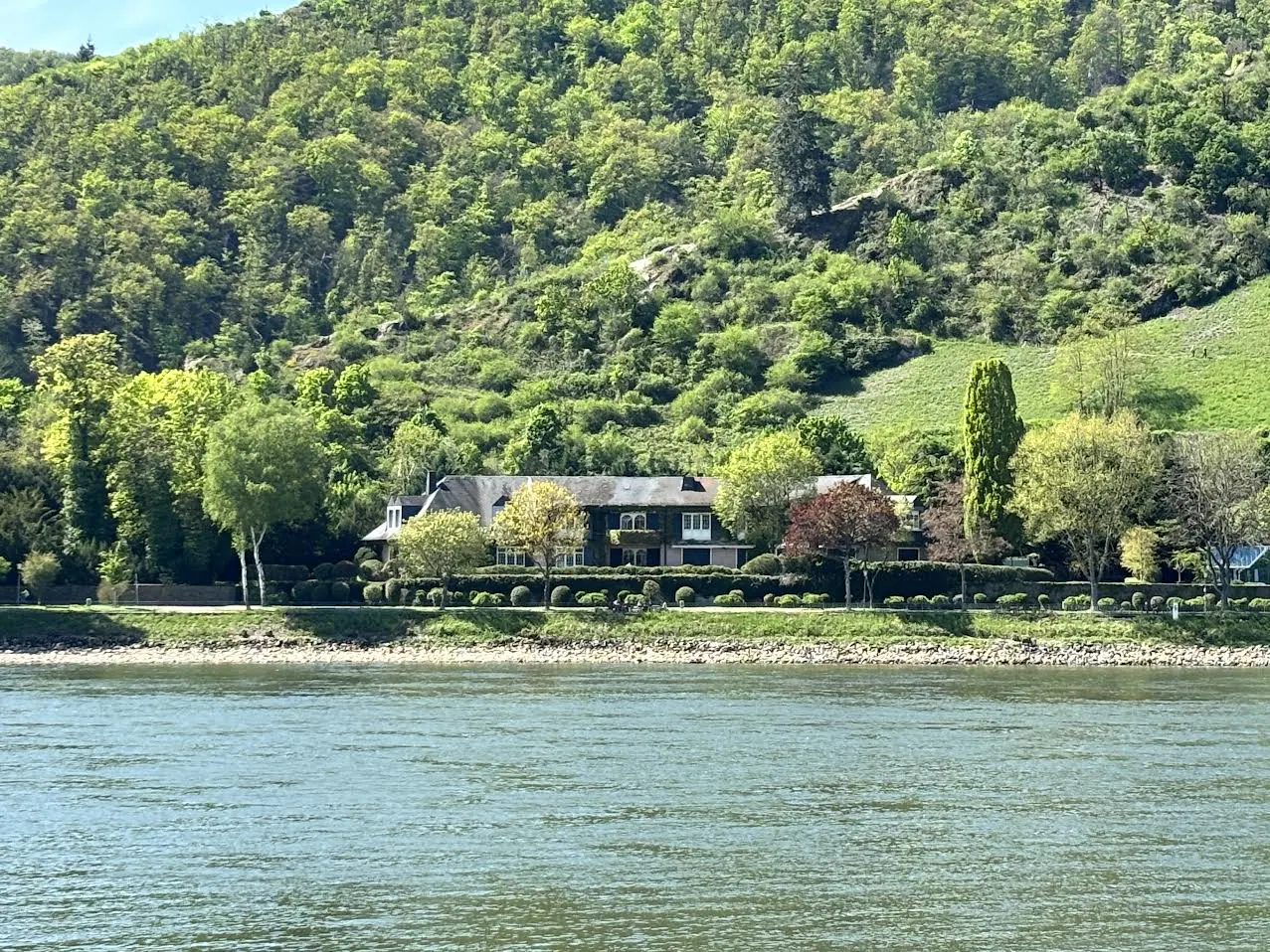 Waterfront property with a large house, green trees, and a lush green hillside in the background.