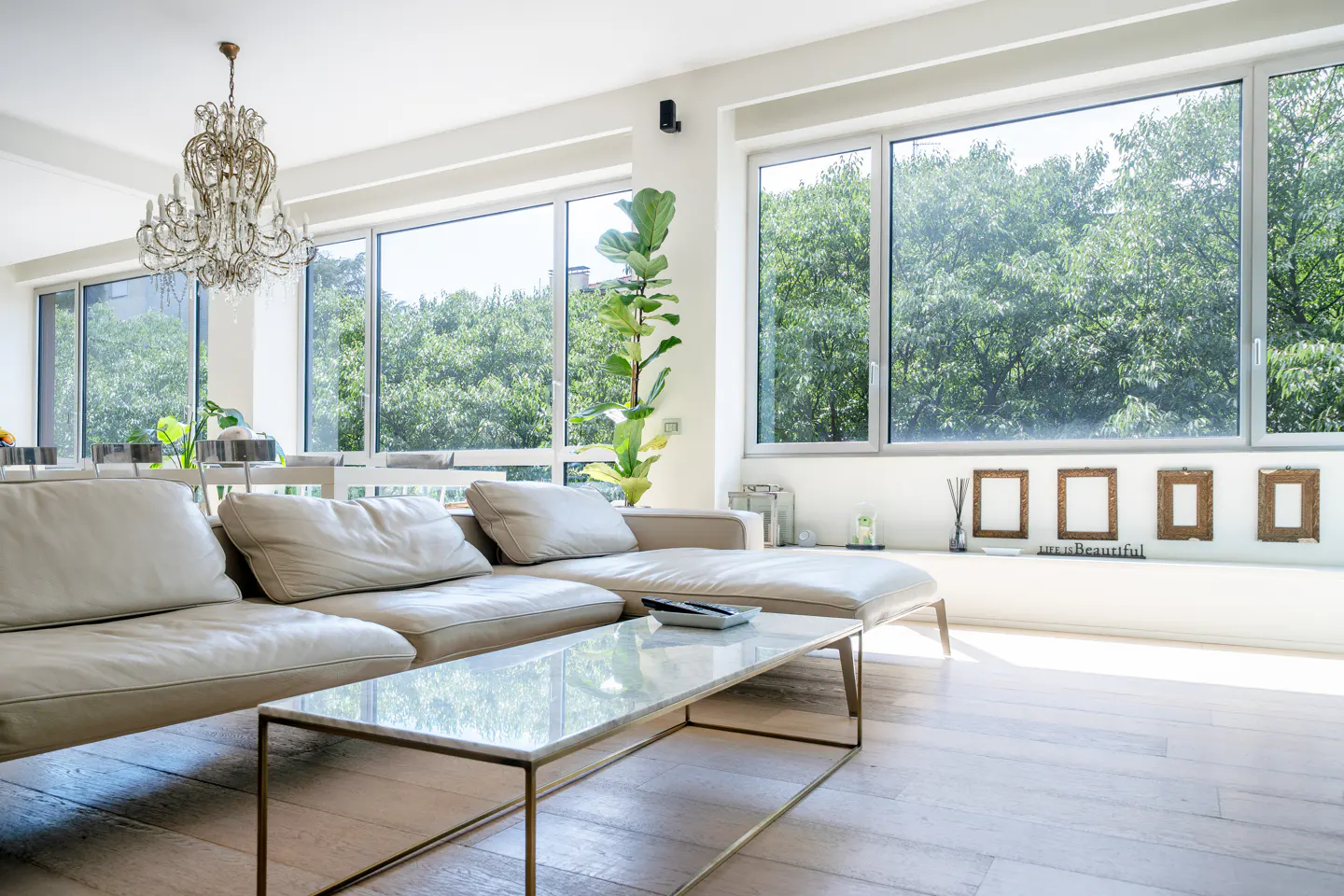 Bright living room with a beige sectional sofa, marble coffee table, crystal chandelier, and large windows with tree views.