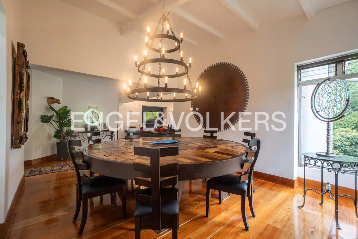 A dining room with a round wooden table, black chairs, and a large chandelier. A decorative saw blade hangs on the wall.