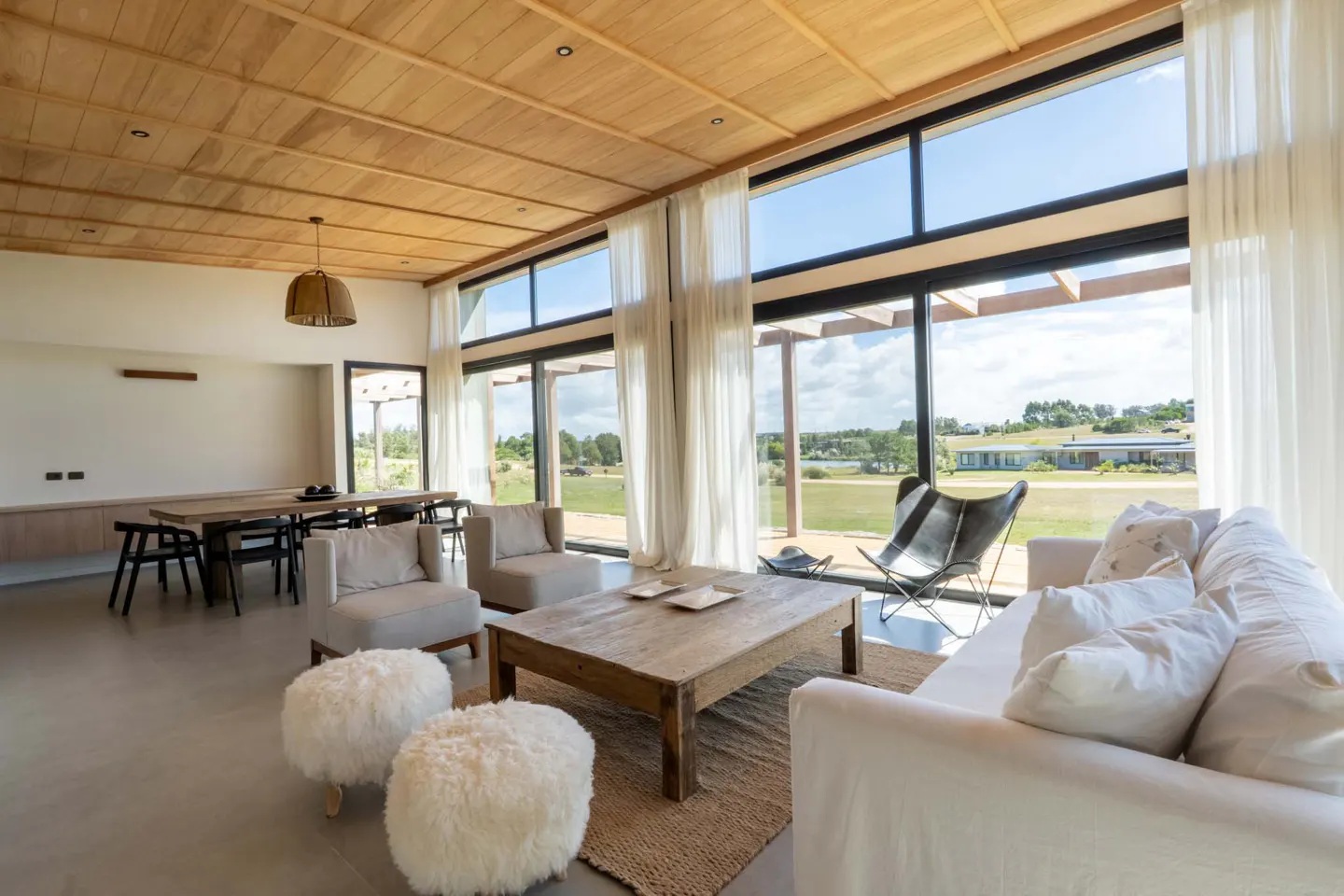 Bright living room with wood ceiling, large windows, and white furniture. A dining table is visible in the background. Outside is a green field.