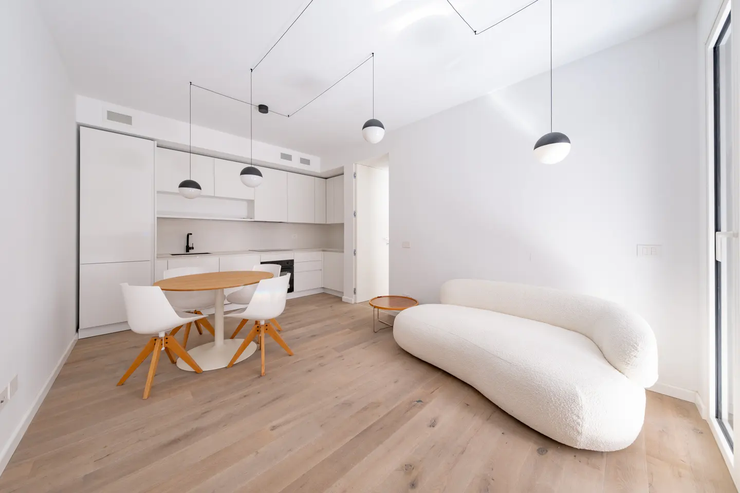 Bright, modern apartment with white walls, wood floors, and open kitchen. Features a round wood table with white chairs and a curved white sofa.