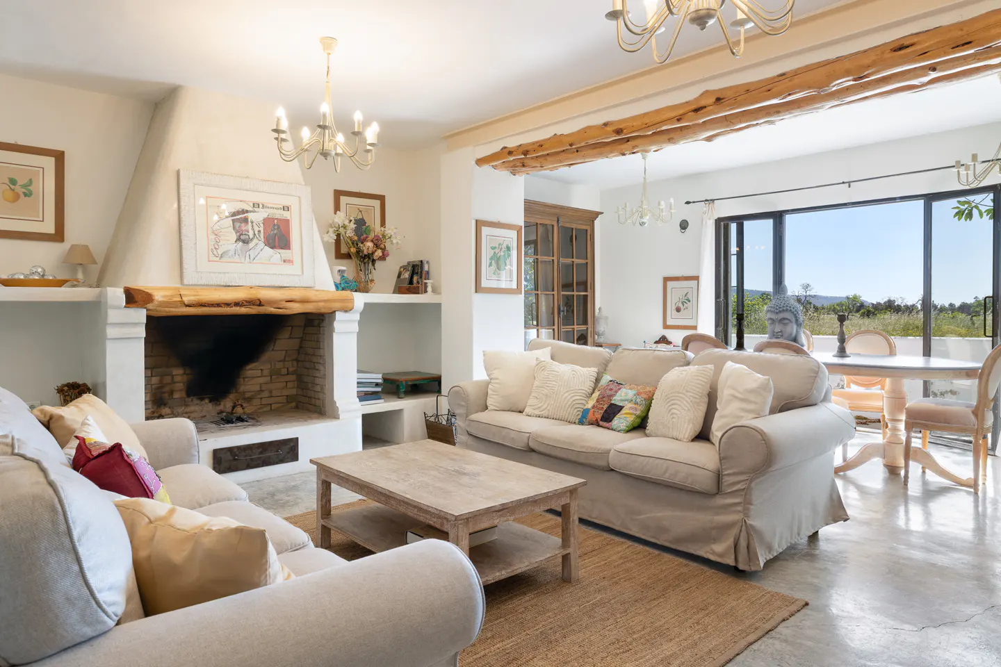 Bright living room with beige sofas, a wooden coffee table, and a fireplace. A dining area is visible through a large opening.