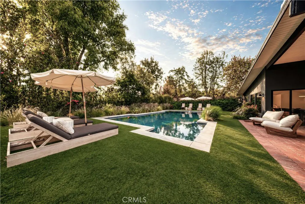 A backyard with a pool, lounge chairs, and umbrellas on a sunny day. The house is dark gray with a red brick patio.