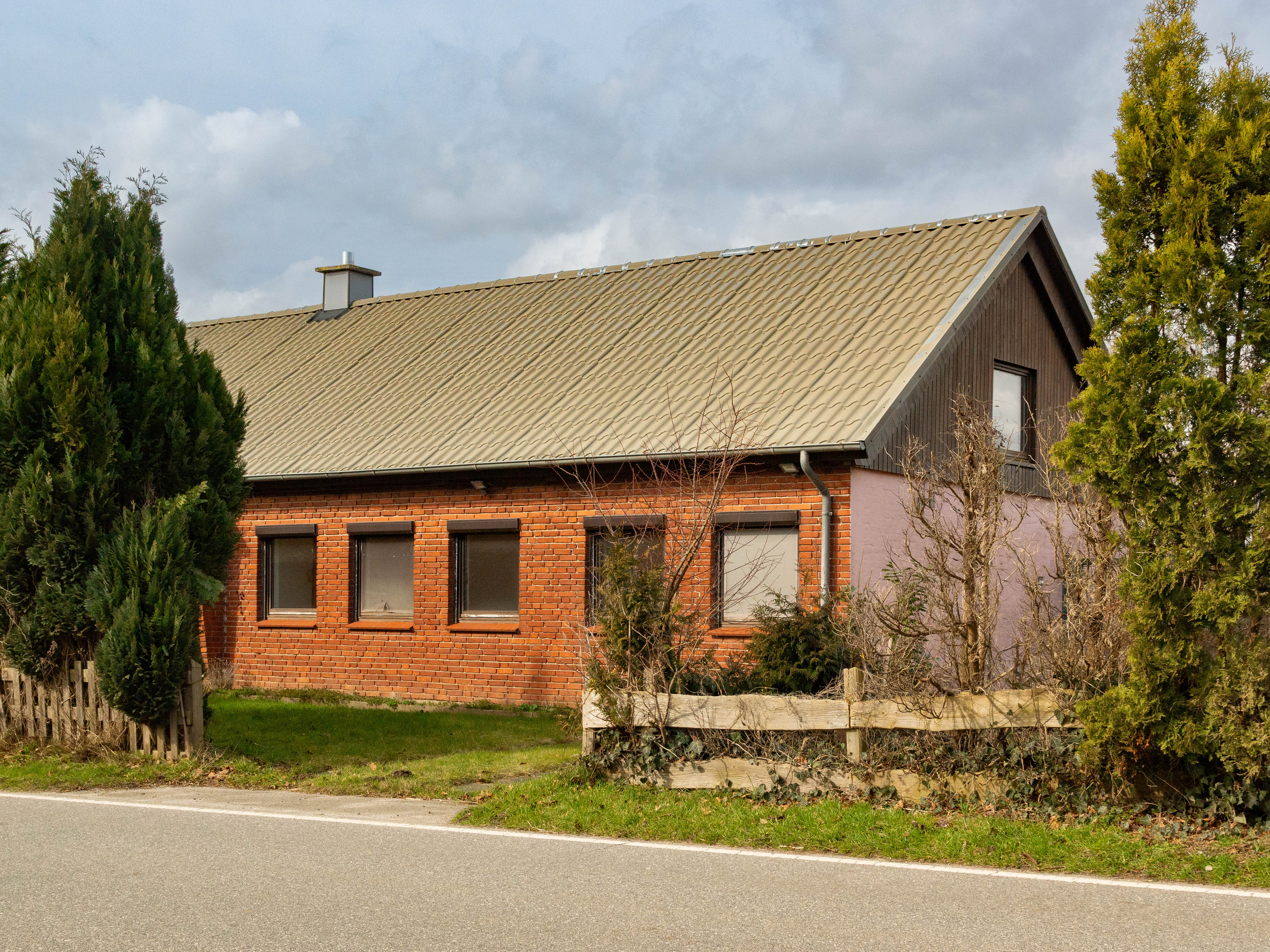 A one-story red brick house with a tan roof and dark trim, surrounded by green trees.