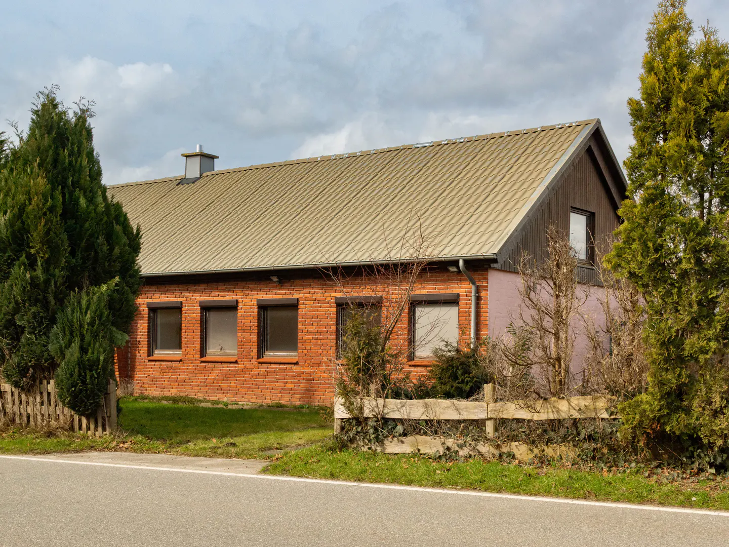 A one-story red brick house with a tan roof and dark trim, surrounded by green trees.