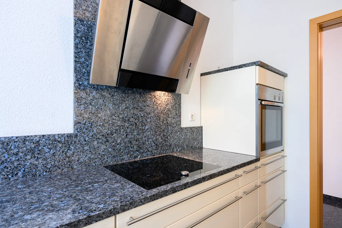 Modern kitchen with granite countertops, stainless steel range hood, and built-in oven. Cream-colored cabinets with silver handles.