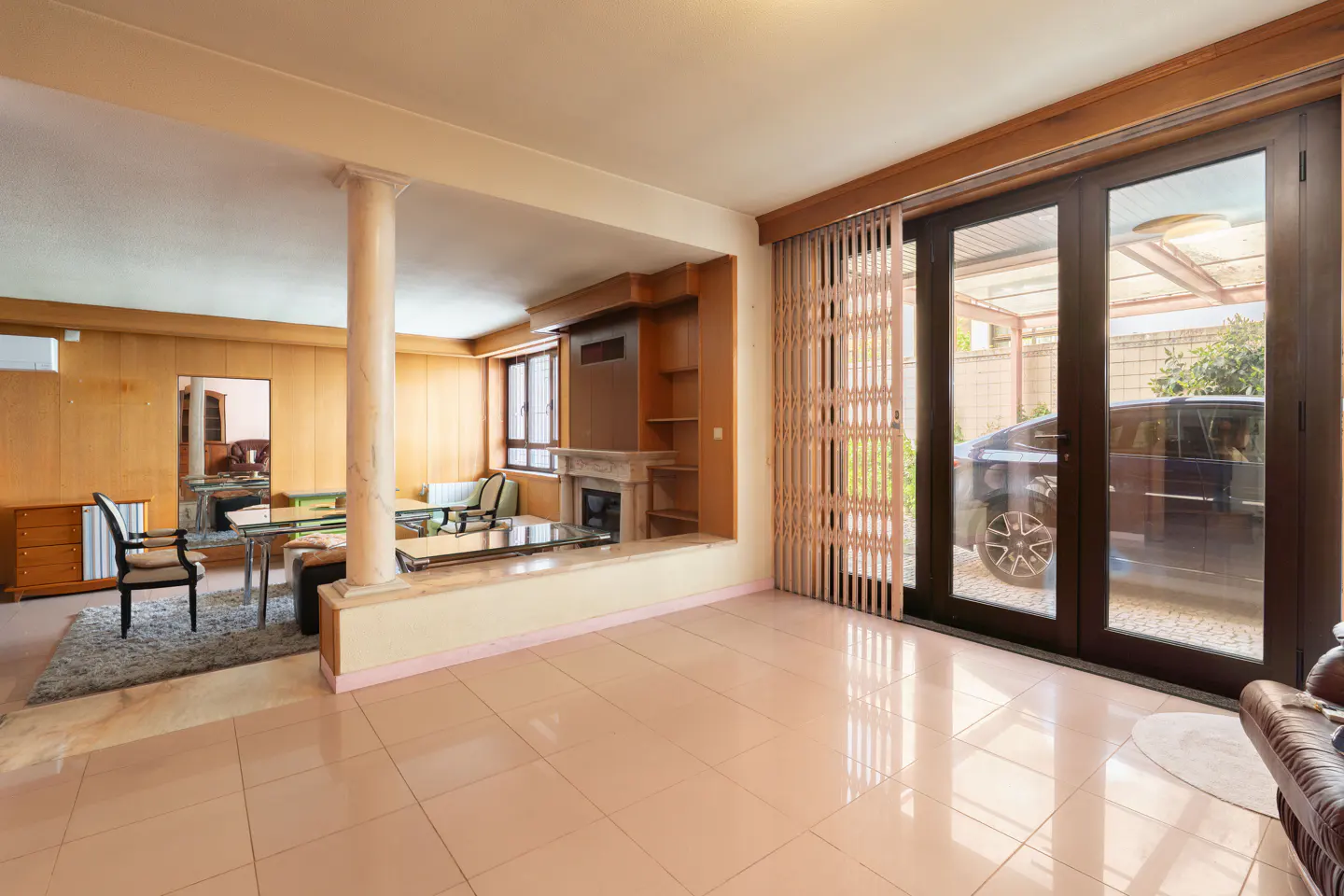 A bright living room with a fireplace, wood paneling, and a large sliding glass door to a carport with a car.