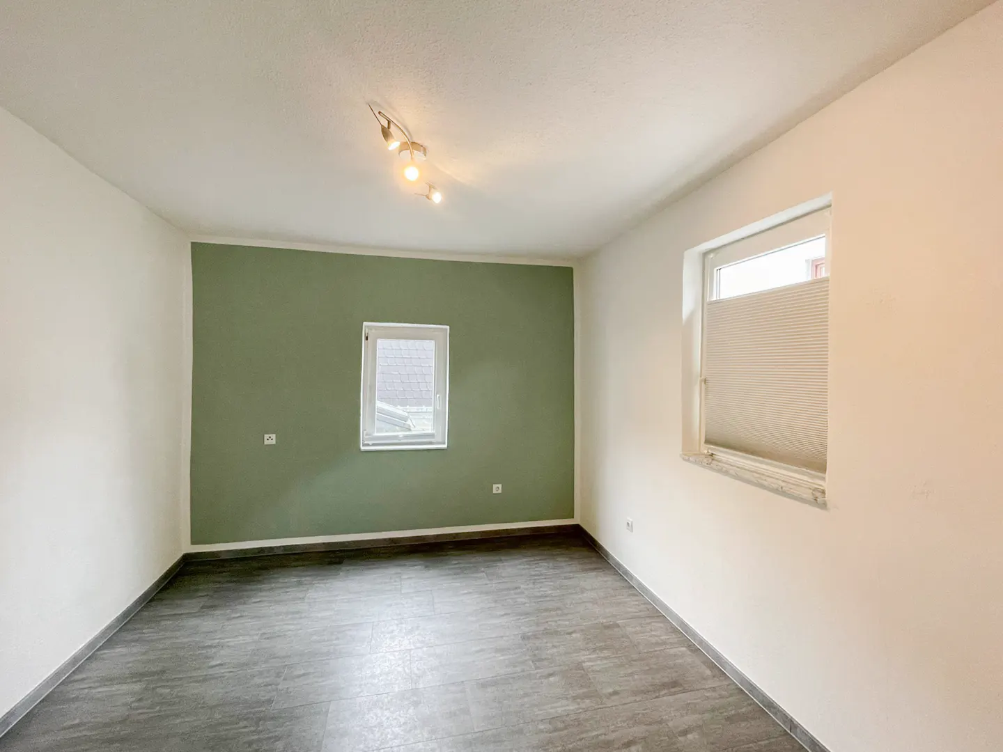 Empty room with gray wood-look floors, white walls, and one green accent wall. Two windows let in natural light.