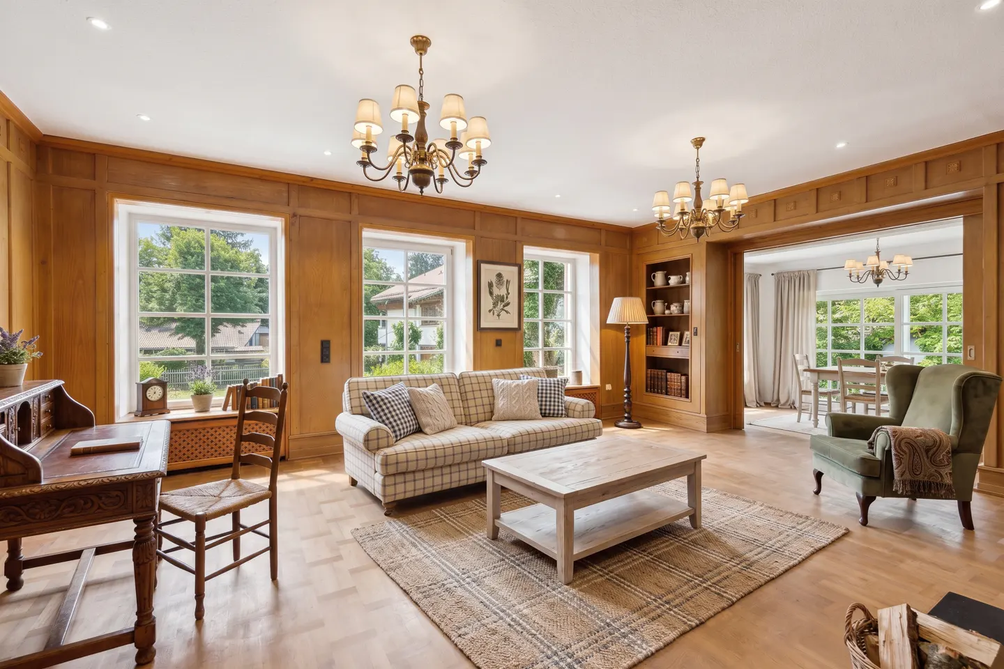 Living room with wood paneling, hardwood floors, and a plaid sofa. Windows overlook a green yard. Chandeliers hang from the ceiling.