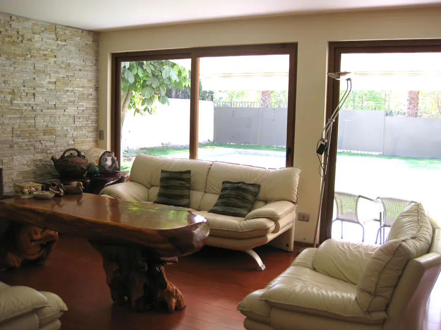 Living room with a stone wall, wood floors, and large windows overlooking a green lawn. A unique wood table and cream-colored sofas furnish the space.
