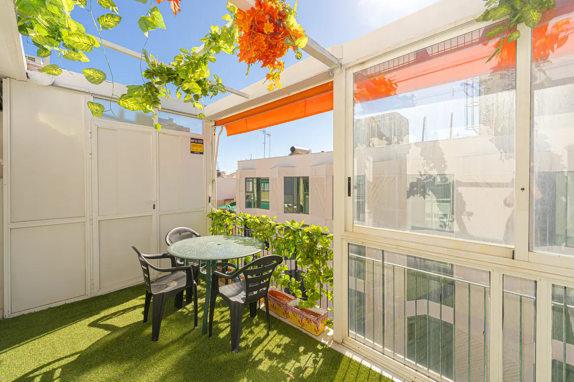 Balcony with artificial grass, a green table, and four black chairs. Green vines and orange flowers decorate the white walls and ceiling.