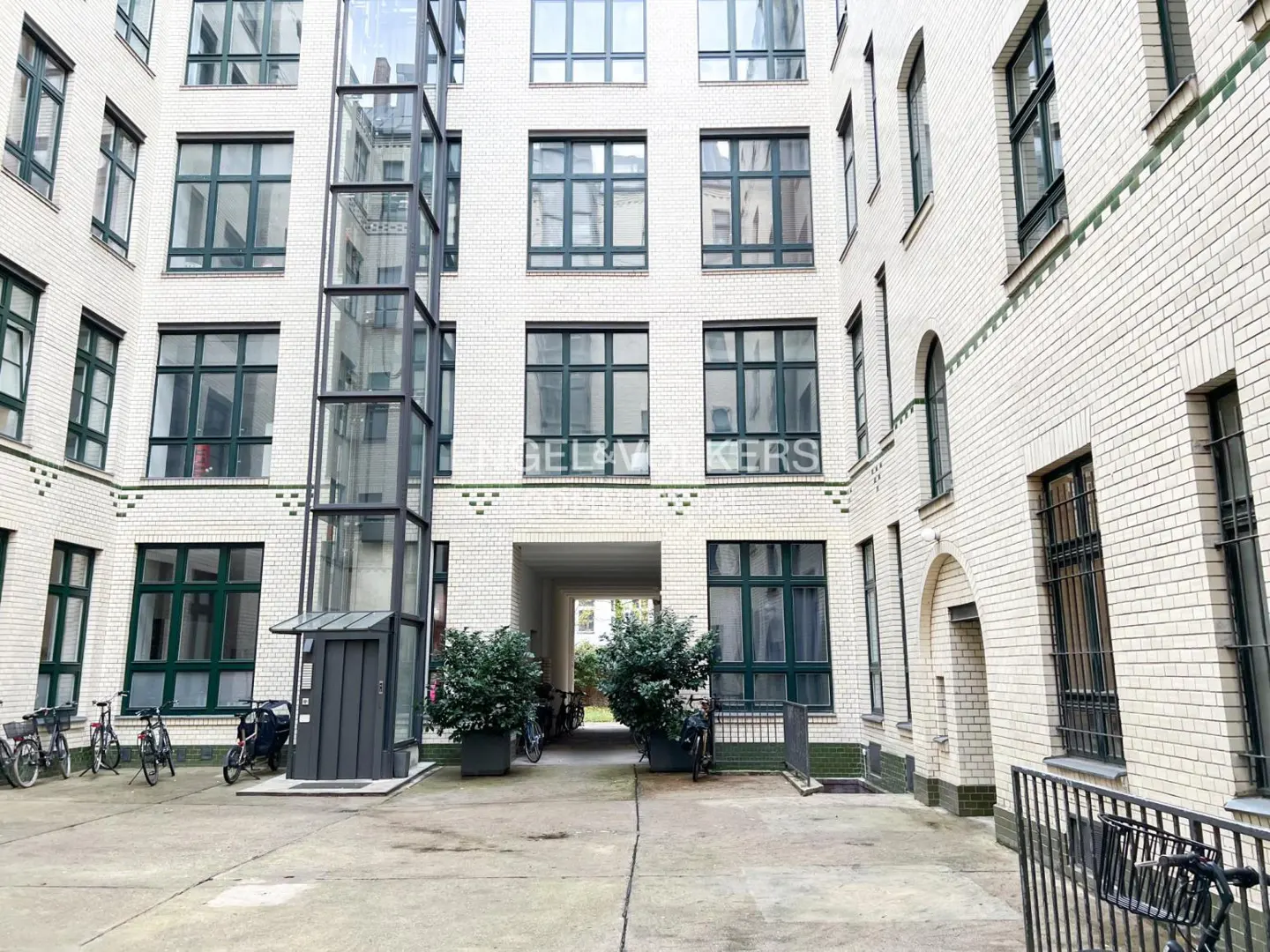 Courtyard view of a white brick building with green window frames and a glass elevator. Bicycles are parked nearby.