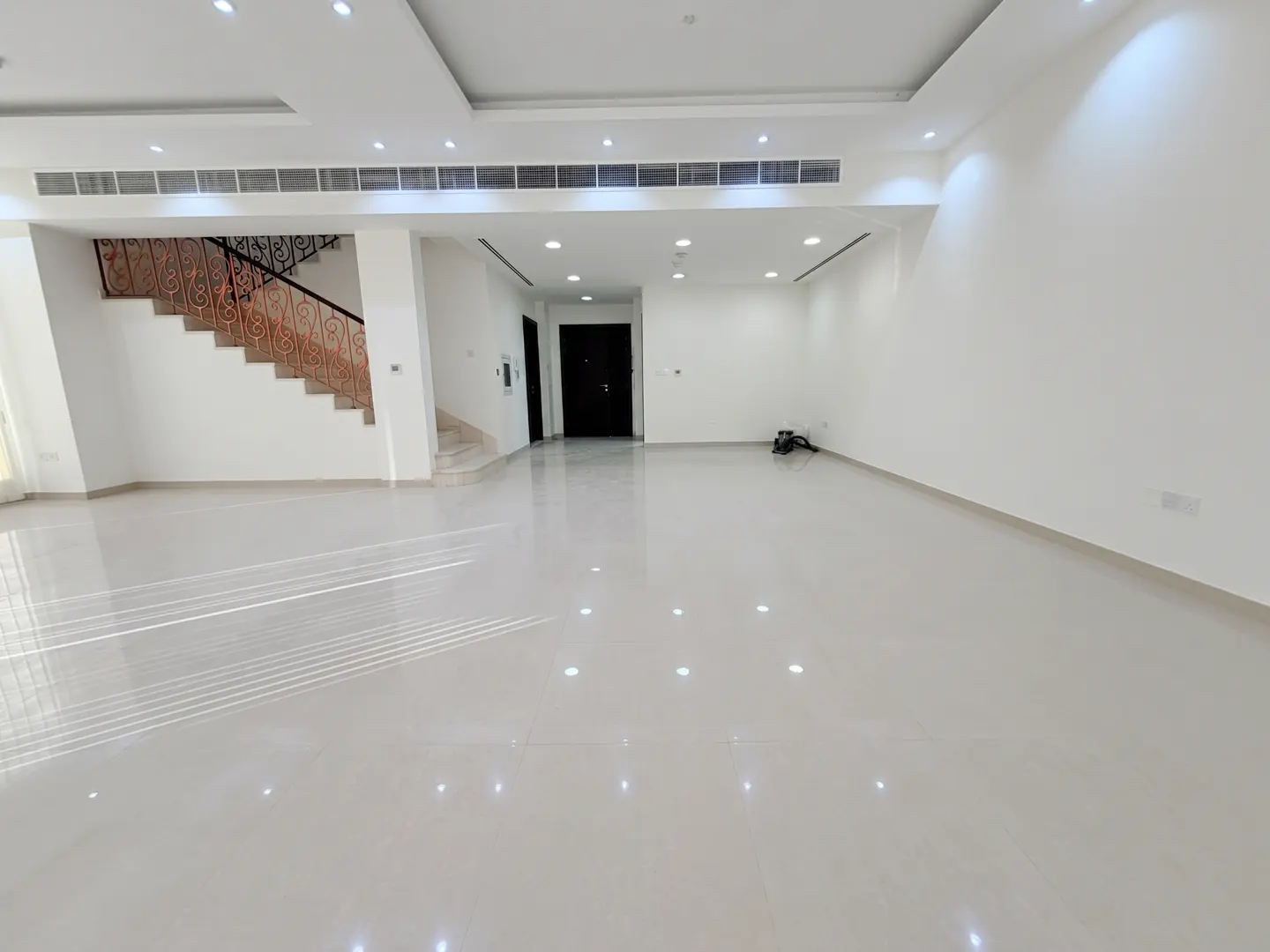 Bright, empty living room with white walls and tile floors. Stairs with decorative railing on the left. Black doors in the back.
