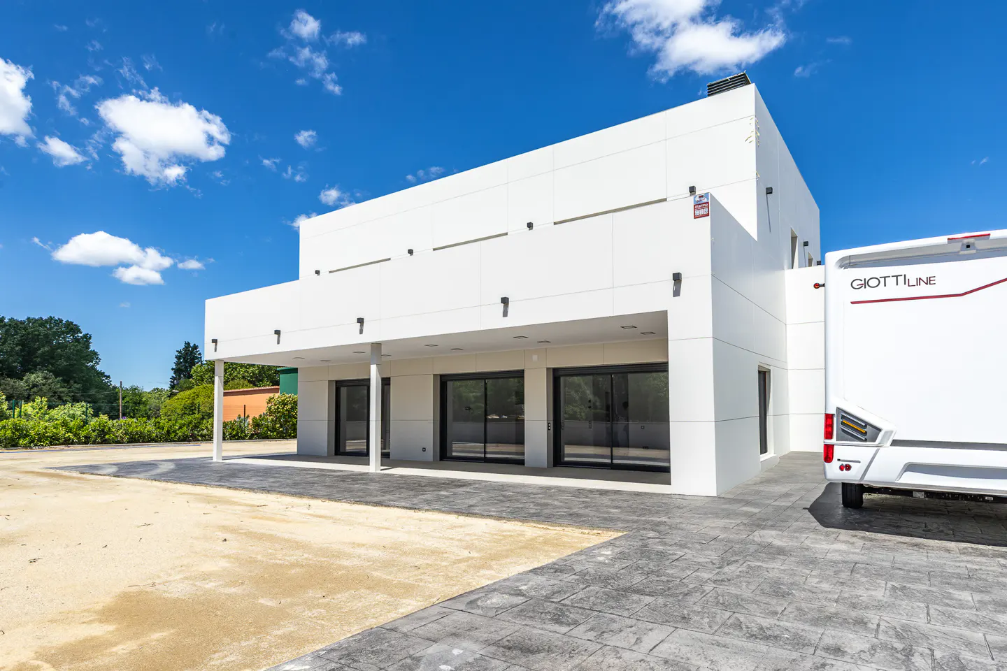 Modern white building with black-framed glass doors, a covered porch, and a parked white Giottiline RV under a blue sky.