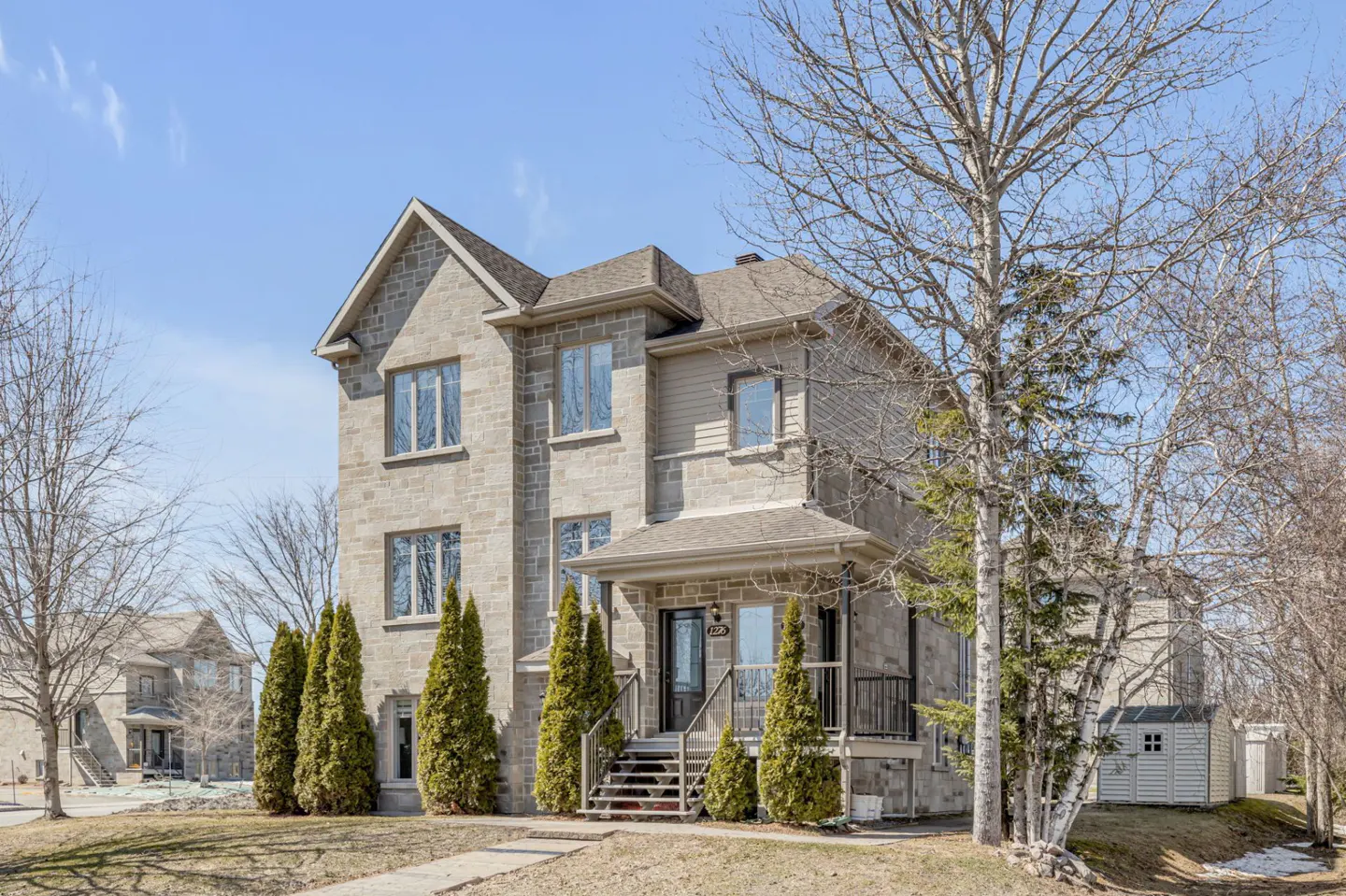 Two-story stone house with a front porch and stairs, surrounded by green trees and a blue sky.
