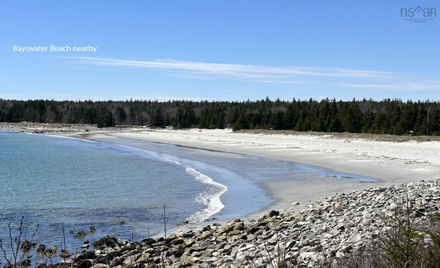 Scenic view of Bayswater Beach with blue water, sandy shore, and a backdrop of green trees under a clear blue sky. Rocky shoreline in the foreground.