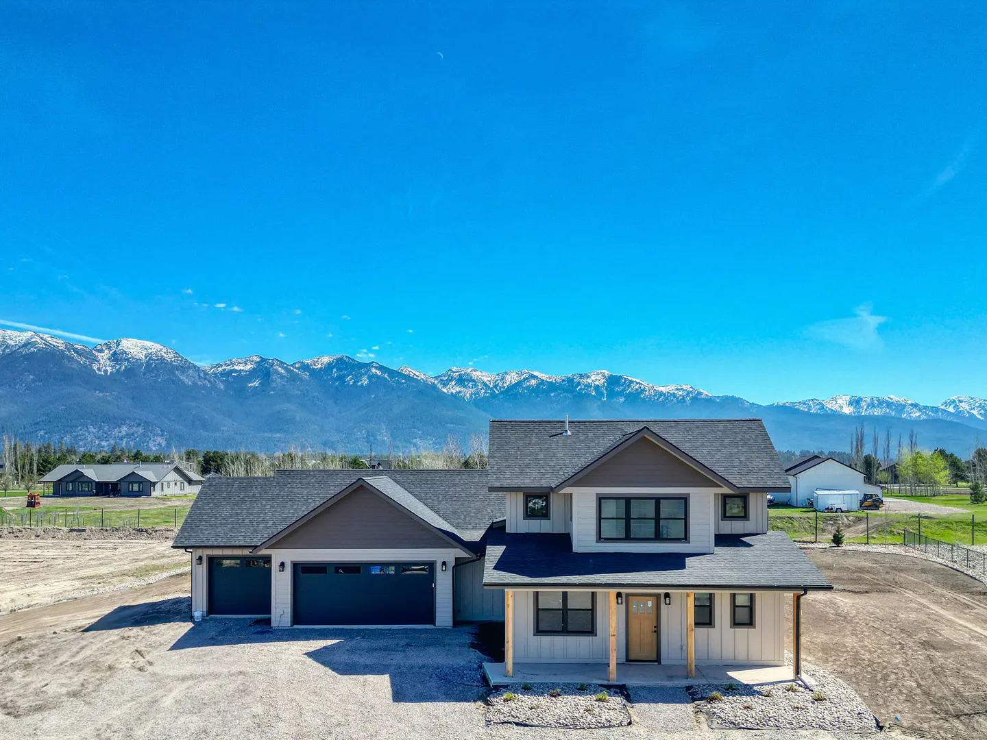 Two-story house with a gray roof, beige siding, and a two-car garage, set against a backdrop of snow-capped mountains and a clear blue sky.