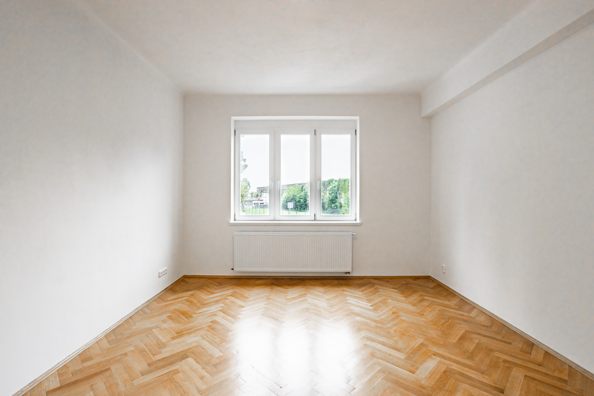 Empty room with white walls, light wood herringbone floors, a central window, and a white radiator underneath.