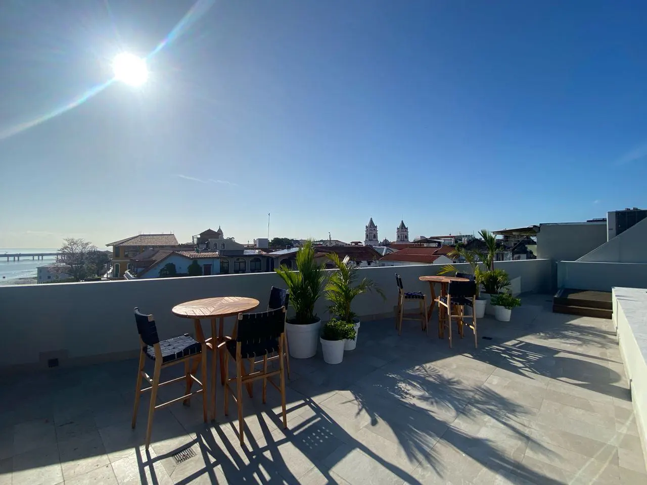 Rooftop patio with two tables, chairs, and potted plants overlooking a city with a clear blue sky and bright sun.